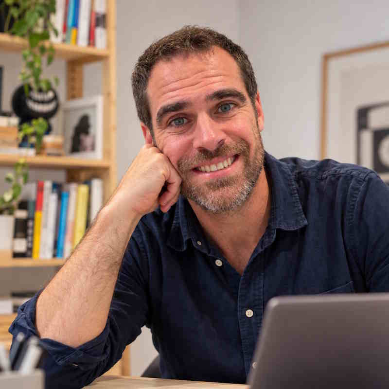 Joan Martín sonriendo en su mesa de trabajo, con una estantería de libros al fondo